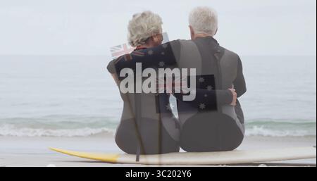Assis couple senior portant des combinaisons et embrassant sur une planche de surf à la rive sablonneuse, avec l'horizon de la mer Banque D'Images