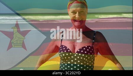 Femme senior souriante en maillot de bain, tenant l'anneau jaune au rivage avec bonnet de bain rouge, lunettes de natation Banque D'Images