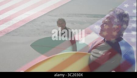 Couple de personnes âgées debout portant des combinaisons tenant des planches de surf sur la plage, avec drapeau recouvrant les vagues Banque D'Images