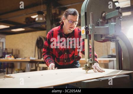 Homme mûr portant chemise en flanelle guidant le contreplaqué à travers la scie à ruban verticale dans l'atelier de bois avec de la sciure de bois Banque D'Images