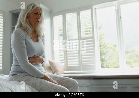 Femme senior serrant l'abdomen assis sur le lit dans la chambre avec volets et oreillers, espace copie Banque D'Images