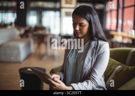 Femme asiatique portant une tenue d'affaires assise sur une chaise vert olive tenant une tablette au salon du café Banque D'Images