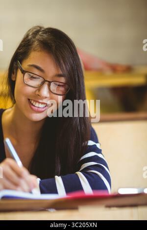 Femme asiatique assise au bureau dans la salle d'étude portant des lunettes écrivant dans un cahier avec stylo argenté Banque D'Images