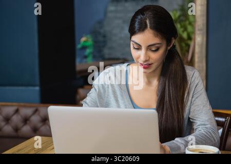 Femme assise à une table de café en bois travaillant sur un ordinateur portable argenté tout en tenant une tasse à café Banque D'Images