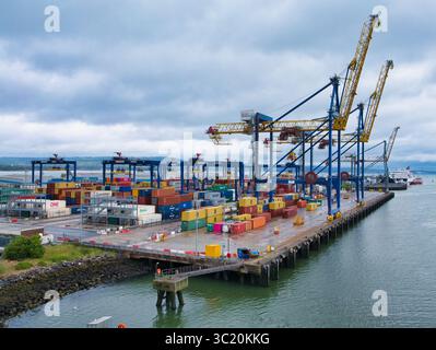 Belfast, Royaume-Uni - 15 juin 2025 : de grandes grues et des conteneurs maritimes empilés bordent les quais animés du port de Belfast sous un ciel nuageux. Banque D'Images