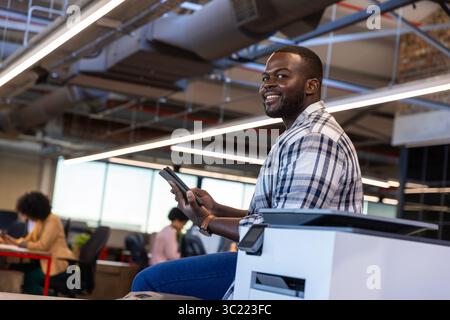 Divers collègues utilisant une tablette, assis sur l'imprimante dans un bureau ouvert avec des lumières LED, espace de copie Banque D'Images