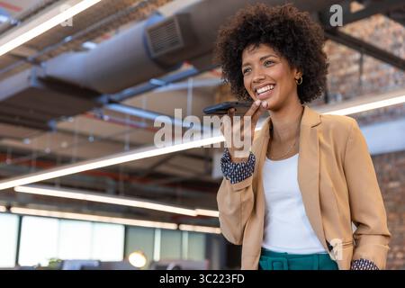 Femme afro-américaine portant blazer parlant dans un smartphone debout dans le bureau, espace de copie Banque D'Images