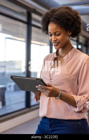 Femme afro-américaine debout dans le couloir du bureau vérifiant la tablette et jetant un coup d'œil à la montre-bracelet Banque D'Images