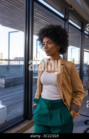 Femme afro-américaine debout près des fenêtres dans le bureau portant blazer, pantalon sarcelle donnant sur la skyline Banque D'Images