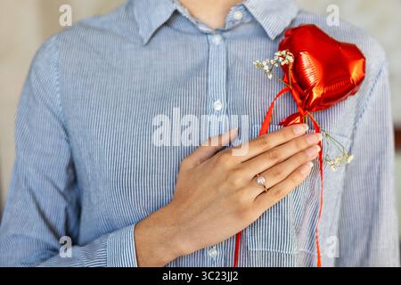 Femme tenant un ballon de coeur avec anneau de fiançailles. Gros plan d'une femme en chemise rayée tenant un ballon coeur rouge et portant une bague de fiançailles Banque D'Images