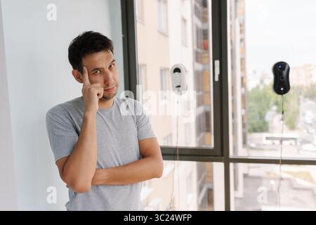 Portrait d'un homme heureux debout à côté d'une grande fenêtre, les bras croisés, observant deux nettoyeurs de vitres robotisés faisant le travail, avec une expression détendue sur le visage Banque D'Images