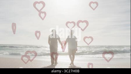 Marchant couple âgé tenant les mains le long de la rive sablonneuse par les vagues de l'océan, avec des superpositions en forme de coeur Banque D'Images