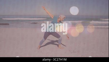Femme senior pratiquant la pose de yoga debout sur la plage de sable, avec haut sarcelle et leggings gris Banque D'Images