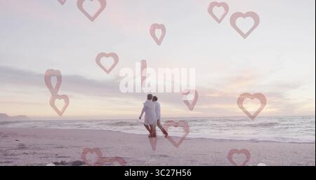 Couple senior marchant portant des vêtements légers bras dans bras sur la plage de sable au coucher du soleil, lueur du soir Banque D'Images