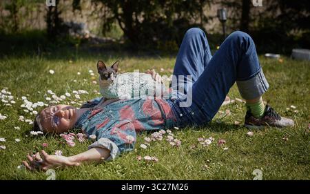 Une femme et son chat Sphynx reposent au soleil sur une pelouse verte entourée de fleurs printanières. Banque D'Images