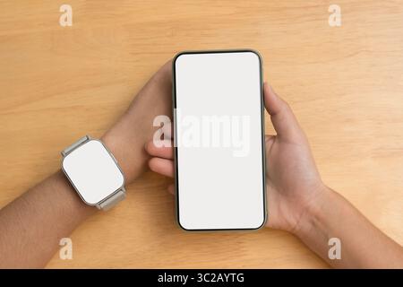 Gros plan d'une personne tenant un smartphone avec un écran blanc vierge et portant une montre intelligente avec un écran vide. Les deux dispositifs placés contre le t en bois Banque D'Images