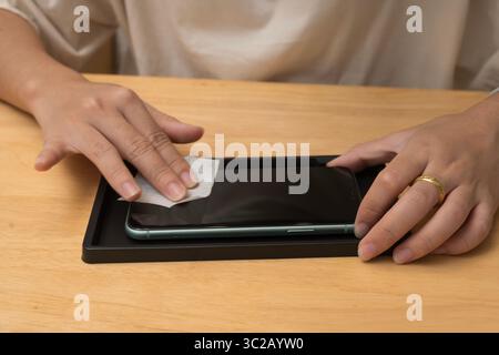 Gros plan des mains essuyant l'écran d'un smartphone avec un chiffon de nettoyage avant d'appliquer un protecteur d'écran. Téléphone repose dans un plateau noir sur une table en bois. P Banque D'Images