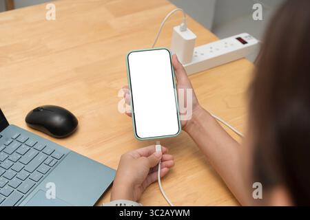 Une personne connecte un smartphone à un chargeur tout en utilisant un ordinateur portable sur une table en bois. Écran vide sur l'appareil mobile. Concept de multitâche, espace de travail, Banque D'Images