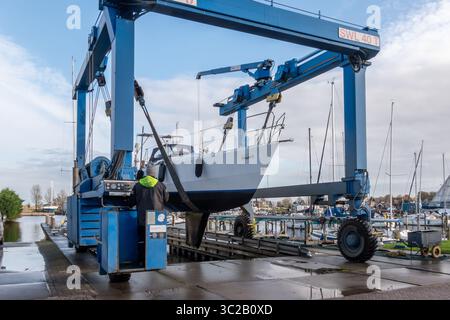 Warns, pays-Bas - 20 novembre 2023 : grue de levage de bateau hissant un voilier hors de l'eau dans une marina animée, avec des ouvriers habilement opératinés Banque D'Images