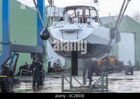 Warns, pays-Bas - 20 novembre 2023 : un ouvrier de chantier naval nettoie la coque d'un voilier avec un nettoyeur haute pression, alors qu'il est soulevé par une grue, entouré Banque D'Images