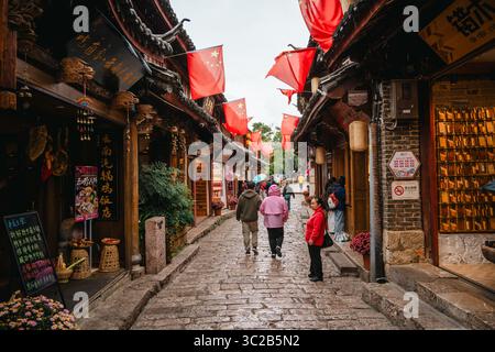 Lijiang, Chine - 23 octobre 2024 : les gens marchent entre les vitrines en bois sous les drapeaux chinois lumineux dans une rue animée de la vieille ville de Lijiang, Yunnan, Chin Banque D'Images