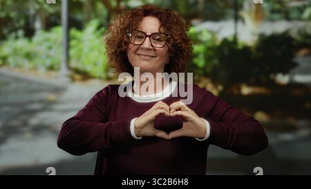 Femme souriant dans une rue de la ville forme une forme de coeur avec ses mains tout en portant des lunettes et un pull Bordeaux, entouré de verdure dans un e extérieur Banque D'Images
