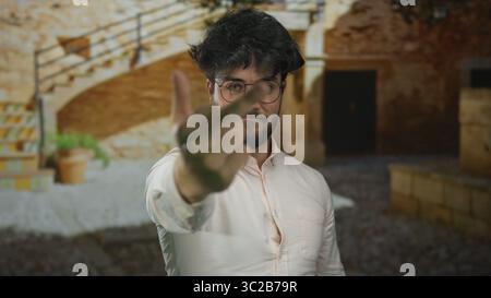 Jeune homme barbu à l'extérieur sur une journée ensoleillée faisant un geste grossier montrant le majeur dans la cour avec des murs de pierre et escalier en arrière-plan Banque D'Images