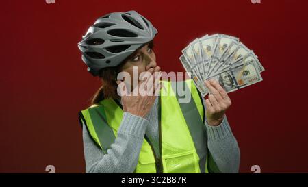 Femme senior portant casque de vélo et gilet de sécurité tient fan de billets de banque américains avec une expression étonnée contre le mur rouge. Banque D'Images