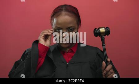 Juge afro-américain en robe noire ajuste les lunettes tout en tenant un marteau en bois dans le studio rouge ; confiance de l'autorité. Banque D'Images