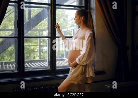 Une femme enceinte se tient près d'une grande fenêtre, berçant doucement sa bosse de bébé. Elle regarde contemplativement dehors tout en étant habillée dans une tenue confortable, cre Banque D'Images