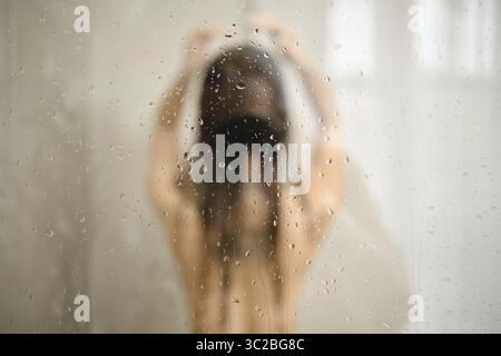 Une femme se tient sous la douche avec les cheveux baissés, profitant d'un moment de paix. L'eau coule le long de son corps, créant une atmosphère sereine avec des gouttelettes Banque D'Images