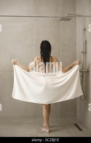 Une femme se tient sous une douche tenant une grande serviette blanche. Des gouttelettes d'eau s'attardent sur ses cheveux mouillés. La toile de fond présente des tuiles neutres, améliorant le calme Banque D'Images