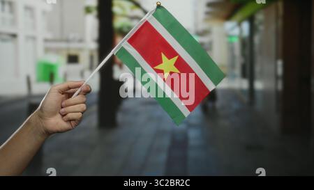 Main tenant le drapeau du suriname sur une rue de la ville à l'extérieur pour signifier la célébration et la fierté d'un homme caucasien au milieu du paysage urbain de la ville. Banque D'Images