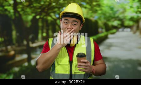 Jeune homme en gilet réfléchissant et casque de sécurité surpris dans le parc extérieur tenant le café. Banque D'Images