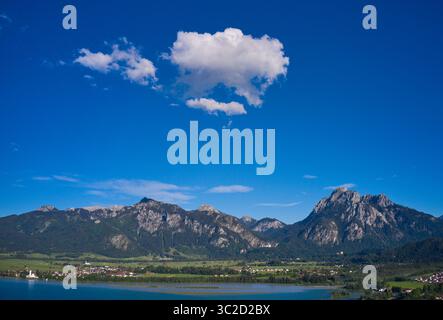Vue aérienne des eaux tranquilles du lac Forggensee reflétant les Alpes majestueuses, nichées au milieu de prairies verdoyantes et de forêts, Füssen, Bayern, Allemagne. Banque D'Images