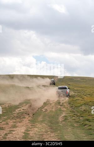 Naryn, Kirghizistan - 12 mai 2024 : deux camionnettes conduisant dans un convoi sur une piste hors route poussiéreuse, avec des sentiers serpentant sur des collines verdoyantes sous un nuage Banque D'Images
