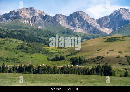 Une vallée de montagne pittoresque avec une ligne de yourtes nichée au milieu de forêts verdoyantes et de collines ondulantes, avec des pics accidentés en arrière-plan Banque D'Images
