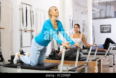 Deux jeunes femmes jeunes adultes et sportives en sport effectuant un exercice Pilates pour le dos sur la machine de reformage de lit dans la salle de gym Banque D'Images
