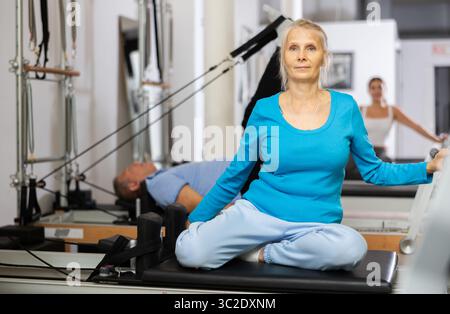 Une femme âgée vêtue de vêtements de sport est assise avec des jambes repliées sur l'équipement du reformeur et s'entraîne à nouveau dans le centre de réadaptation Banque D'Images