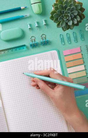 La main de la femme écrit dans un cahier avec de la papeterie turquoise sur un fond de menthe Banque D'Images