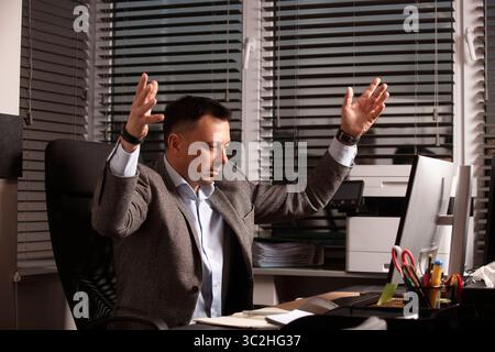 Homme d'affaires bouleversé lève les mains dans la frustration tout en travaillant à un bureau encombré. Symbole d'épuisement professionnel, de stress professionnel ou de démission. Lieu de travail. Banque D'Images