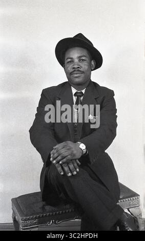 Années 1950, historique, à l'intérieur d'un studio photographique... un gentleman afro-caribéen habillé en costume & cravate & chapeau assis sur un tabouret pour sa photo, Angleterre, Royaume-Uni. À cette époque, avoir votre photographie prise dans un studio, dans un cadre formel était populaire, car les appareils photo 35 mm moins chers et les laboratoires photo pour traiter le film et faire des tirages ne sont pas venus avant les années 1960 Alors à cette époque, pour des services photographiques de qualité, vous alliez chez un photographe professionnel dans un studio. Banque D'Images