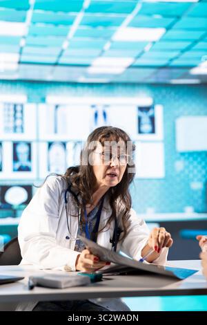 Vieille femme médecin évaluant les rapports de diagnostic au patient dans l'armoire, en examinant les résultats de test de balayage de rayons X après consultation professionnelle. Médecin généraliste offrant des conseils professionnels. Banque D'Images