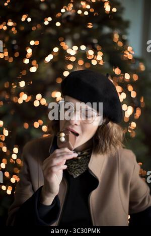 Mignonne jeune femme dans la tenue d'hiver, tout en appréciant mordre dans les gâteries sucrées de vacances chocolat devant l'arbre de Noël. Drôle de femme à la mode profiter Banque D'Images