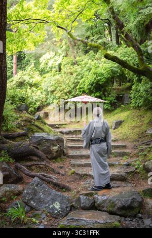 Un homme âgé dans un kimono traditionnel tenant un parapluie wagasa sur des marches en pierre moussue dans une forêt luxuriante. Kyoto, Japon. Banque D'Images