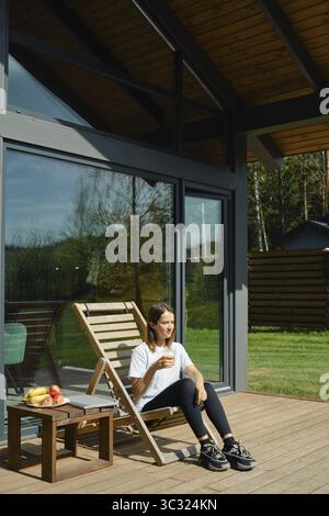 Une femme s'assoit confortablement dans une chaise longue en bois sur une terrasse spacieuse, profitant d'une boisson rafraîchissante Banque D'Images