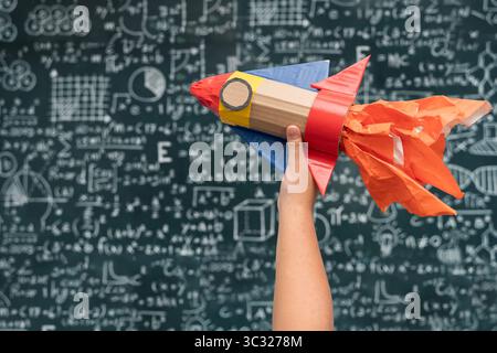 Une main d'enfant tenant une fusée jouet colorée avec des flammes oranges devant un tableau plein d'équations scientifiques et mathématiques, symbolisant le cr Banque D'Images