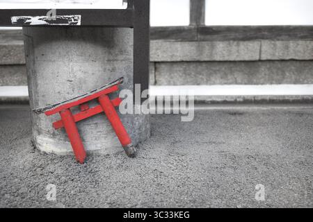 Un petit portail torii rouge cassé s'appuie contre un pilier en béton sur une surface d'asphalte gris. Banque D'Images