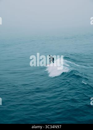 Une figure de surfeur floue solitaire perdue parmi les vagues dans un paysage marin bleu avec du mouvement Banque D'Images