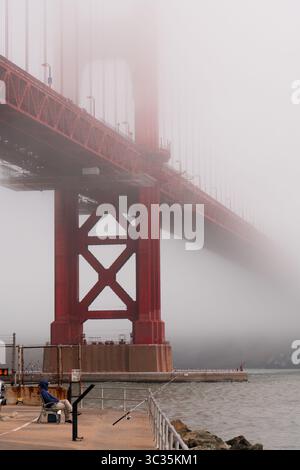Golden Gate Bridge, un pêcheur solitaire assis sur le quai en contrebas près de Fort point. Brouillard estival typique, San Francisco, États-Unis Banque D'Images